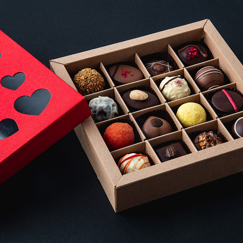 Offene Geschenkbox mit 16 bunten handgemachten Pralinen, roter Herzdeckel daneben – liebevolles Schokoladenpräsent.
